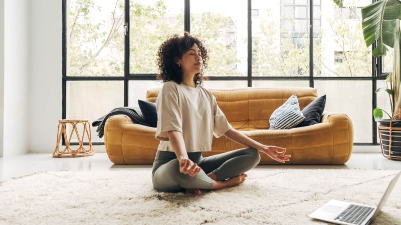 A woman with curly hair sits cross-legged in a meditation pose on a soft rug, in front of a mustard-yellow sofa, inside a bright modern living room with large windows and indoor plants.